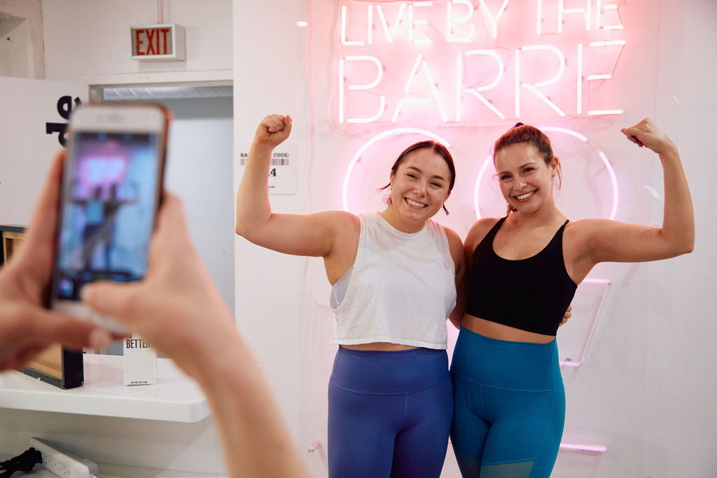 Two women gal pals celebrating their bachelorette party at The Barre Code in River North in Chicago. Post barre / pilates fitness class, these friends glow and hold up flexed biceps in front of The Barre Code's custom mural by Lauren Asta.